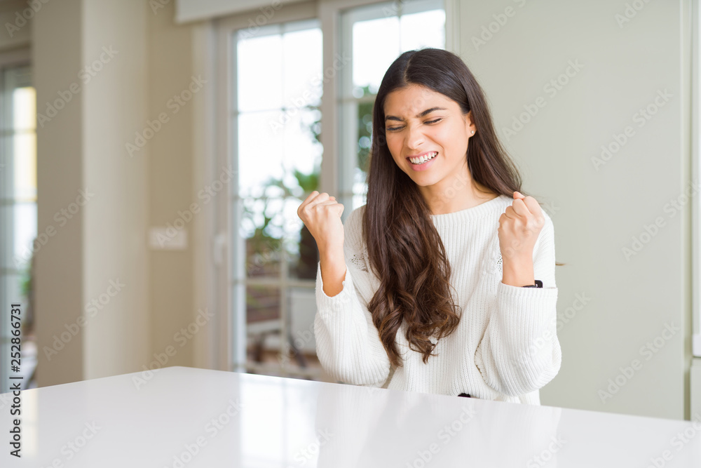 Young beautiful woman at home on white table very happy and excited doing winner gesture with arms raised, smiling and screaming for success. Celebration concept. Young beautiful woman at home on white table very happy and excited doing winner gesture with arms raised, smiling and screaming for success. Celebration concept.