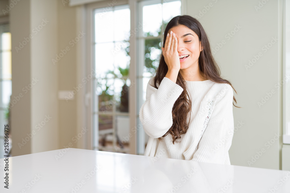 Young beautiful woman at home on white table covering one eye with hand with confident smile on face and surprise emotion. Young beautiful woman at home on white table covering one eye with hand with confident smile on face and surprise emotion.