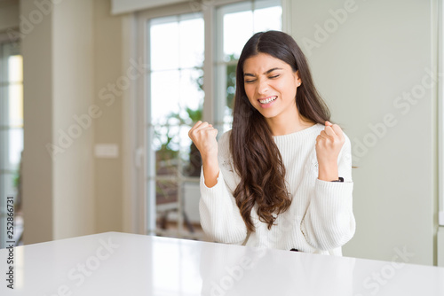 Young beautiful woman at home on white table very happy and excited doing winner gesture with arms raised, smiling and screaming for success. Celebration concept. Free Download Young beautiful woman at home on white table very happy and excited doing winner gesture with arms raised, smiling and screaming for success. Celebration concept. - GFXLoad