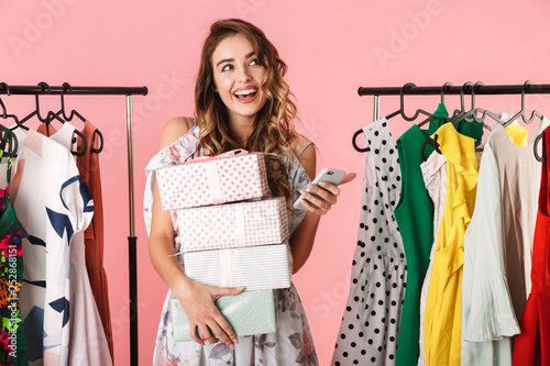 Photo of young woman with purchase standing in store near clothes rack and using mobile phone
