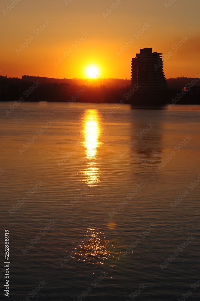 The bright yellow sun sets over the horizon against the black silhouette of the city and a lonely large building above the river.