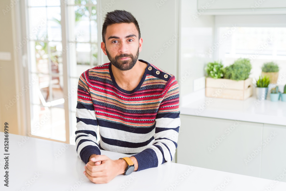 Handsome hispanic man wearing stripes sweater at home with serious expression on face. Simple and natural looking at the camera.