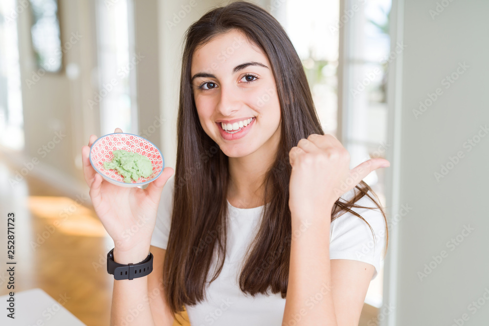 Beautiful young woman holding spicy asian wasabi pointing and showing with thumb up to the side with happy face smiling Beautiful young woman holding spicy asian wasabi pointing and showing with thumb up to the side with happy face smiling