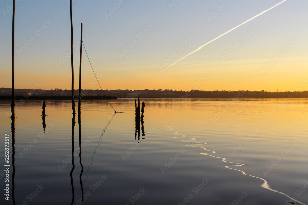 River landscape, with sunset colors, plane trail and wooden sticks reflection