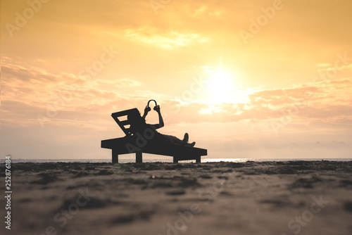 Girl in headphones is sitting on the sun lounger listening to music on the beach at sunset. Summer concept.