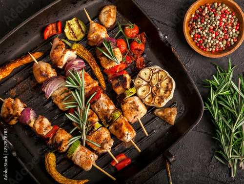 Grilled chicken skewers with spices and vegetables in a pan on black background