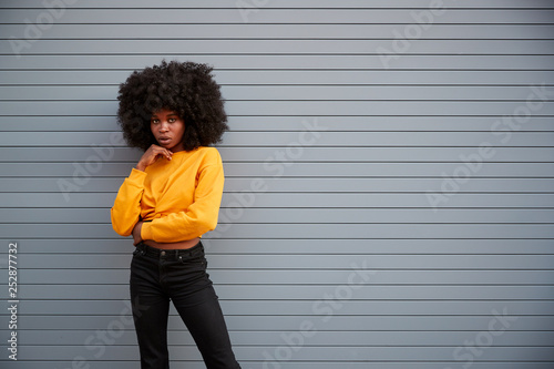 Hip young black woman standing against grey security shutters, head on hand, looking to camera