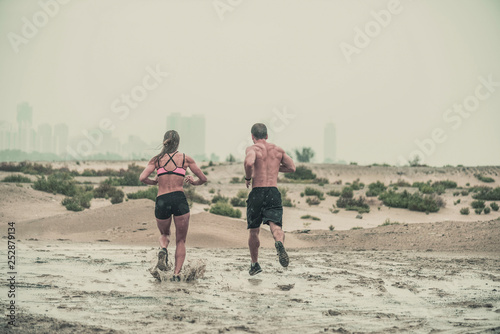 Rear view of muscular male and female athlete covered in mud running down a rough terrain with a desert background in an extreme sport race with grungy textured finish