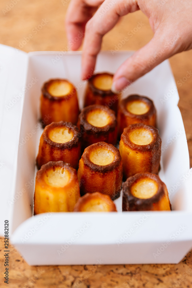 Female hand picking fresh baked Canelés inside white paper box. A small French pastry flavored with rum and vanilla with a soft and tender custard center and a dark, thick caramelized crust.