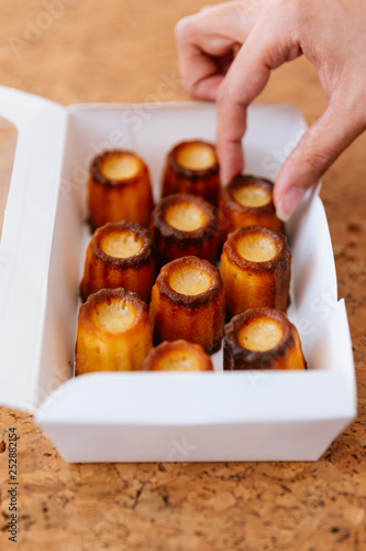 Female hand picking fresh baked Canelés inside white paper box. A small French pastry flavored with rum and vanilla with a soft and tender custard center and a dark, thick caramelized crust. photo