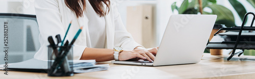 cropped view of businesswoman sitting at table with laptop and typing in office