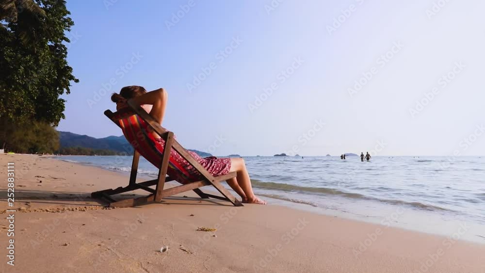 Young woman resting on the beach sitting in deck chair bare foot in the waves, warm summer vacation holidays sea landscape, relaxing outdoor lifestyle