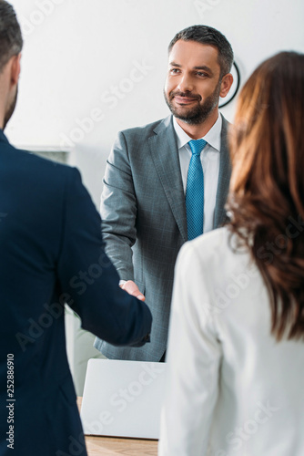 selective focus of handsome advisor in suit shaking hands with investors at office photo