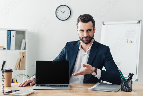 handsome advisor in suit pointing with fingers at laptop with blank screen at office