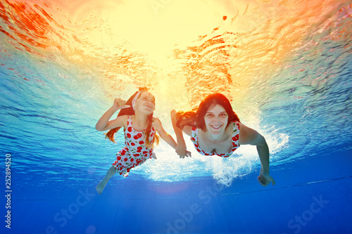 Happy mother and daughter swim under the water holding hands against the bright orange sunset in the same dresses. Portrait. Shooting underwater. Horizontal orientation of the image