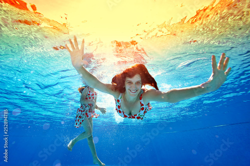Happy family swimming and playing underwater in the pool at sunset. Mom looks at the camera and smiles, spreading her hands to the sides. Portrait. Shooting underwater