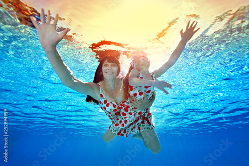 Happy mom and daughter swim hugging underwater in the pool at sunset, spreading his hands to the sides. Mom looks at the camera and smiles. Portrait. Shooting underwater. Bottom view © alexbard