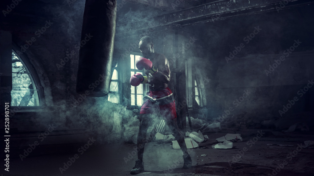 Hands of boxer over dark gym background. Strength, attack and motion concept. Fit african american model in movement. Naked muscular athlete in red gloves. Sporty man during boxing