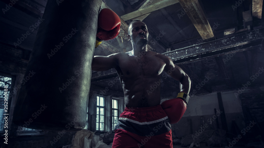 Hands of boxer over dark gym background. Strength, attack and motion concept. Fit african american model in movement. Naked muscular athlete in red gloves. Sporty man during boxing