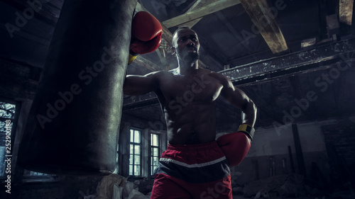 Hands of boxer over dark gym background. Strength  attack and motion concept. Fit african american model in movement. Naked muscular athlete in red gloves. Sporty man during boxing