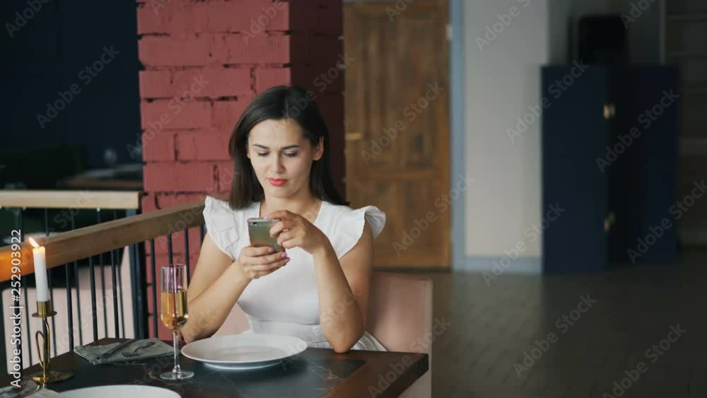 Attractive young woman in beautiful dress is waiting for her boyfriend in restaurant sitting at table alone and using smartphone checking time. Dating and technology concept. Attractive young woman in beautiful dress is waiting for her boyfriend in restaurant sitting at table alone and using smartphone checking time. Dating and technology concept.