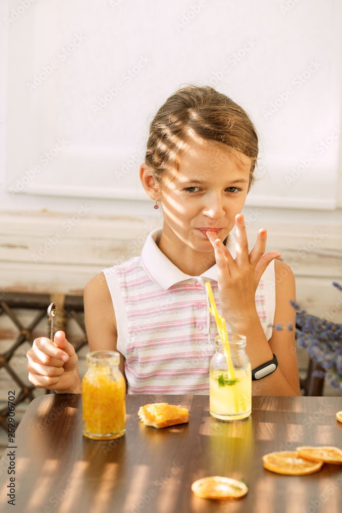 Happy little girl of school age enjoying healthy breakfast eating sandwich and fruits and drinking orange juice sitting at bright sunny terrace Happy little girl of school age enjoying healthy breakfast eating sandwich and fruits and drinking orange juice sitting at bright sunny terrace