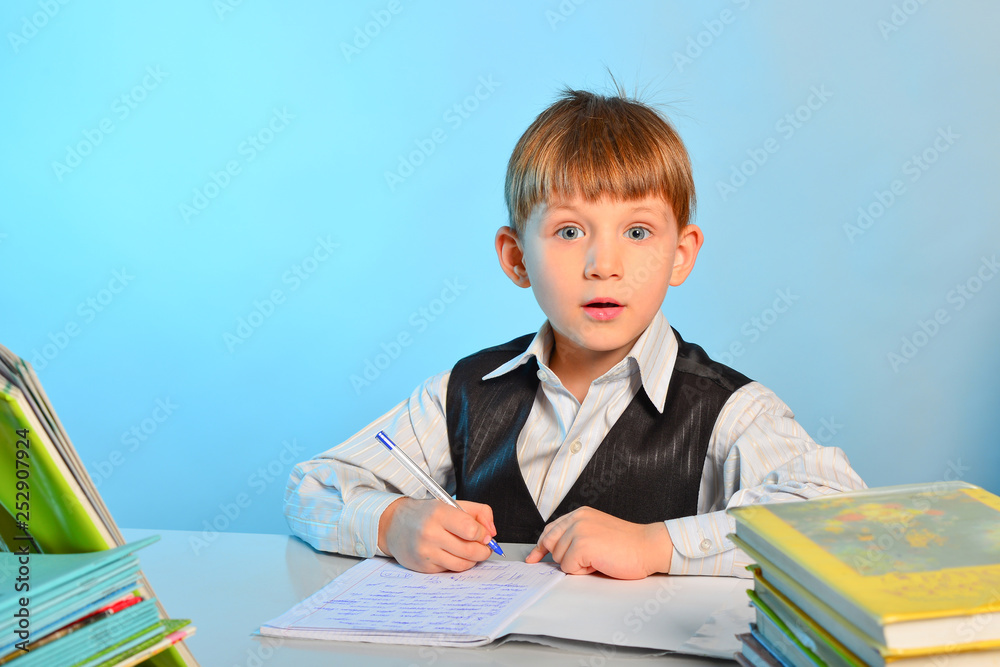 Surprised little schoolboy among notebooks and textbooks at his desk at school in the classroom Surprised little schoolboy among notebooks and textbooks at his desk at school in the classroom
