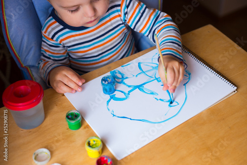 Left hand toddler drawing brush with gouache paint on paper in an album with several colorful brushes near it. photo