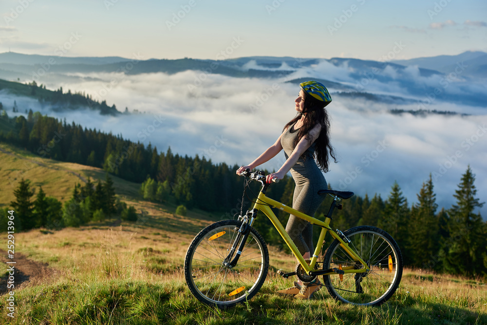 Young female rider standing with yellow bicycle in the mountains, wearing helmet, enjoying valley view on summer morning. Foggy mountains, forests on the blurred background. lifestyle concept
