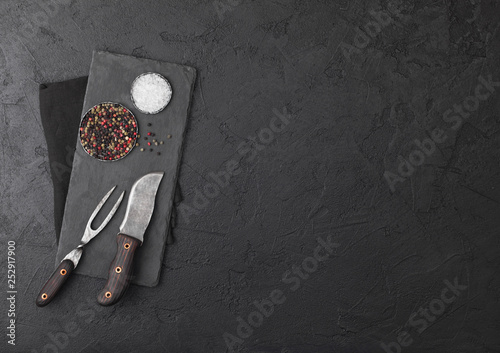 Vintage meat knife and fork with stone chopping board and black table background. Butcher utensils. Salt and pepper.