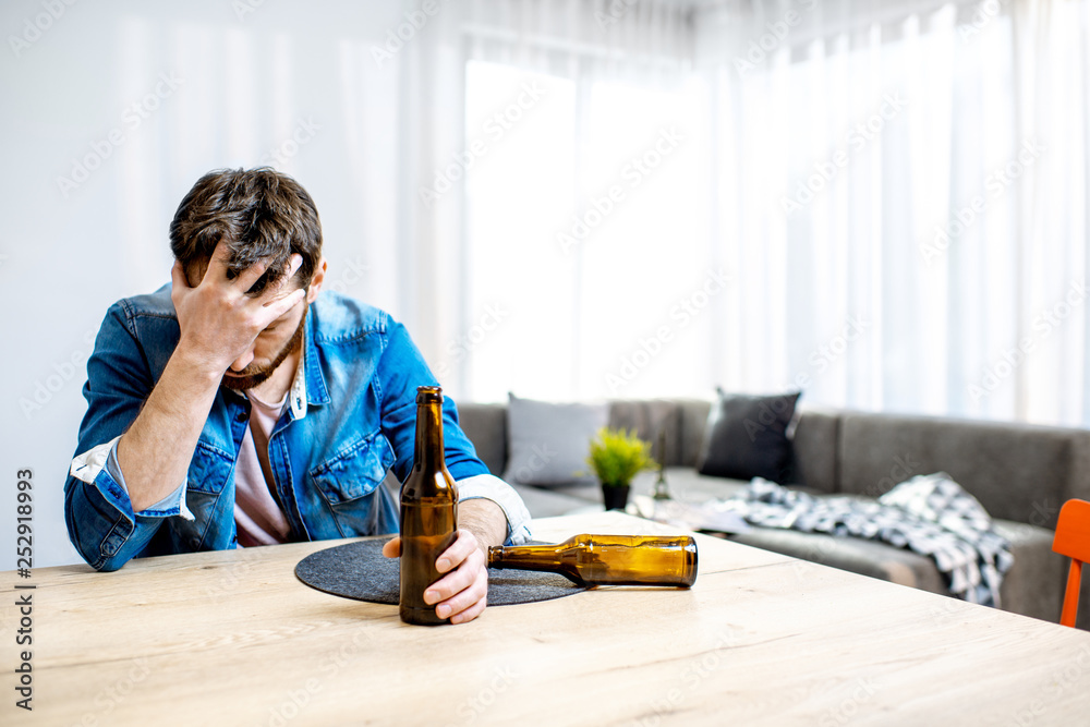 Drunk male alcoholic sitting with bottles feeling depressed and suffering from head ache at home Drunk male alcoholic sitting with bottles feeling depressed and suffering from head ache at home