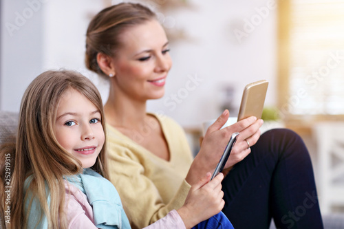 Young mom and her little daughter using smartphone and sitting on sofa at home