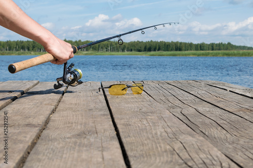 hand puts a fishing rod with a reel next to sun glasses on a wooden pier