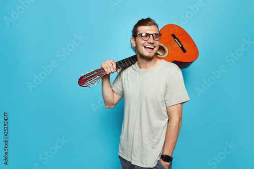 Portrait of young hipster man with acoustic guitar on shoulder smiling and looking away while standing on bright blue background.