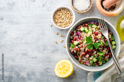 Healthy pearl barley salad with beans, cucumbers, red onion, sunflower seeds, pomegranate and parsley in bowl on concrete background. Top view. Copy space.