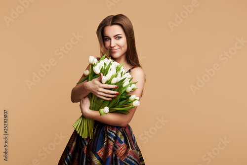 Wow effect. Wonderful flowers on Womens Day. Tender smiling brunet woman poses with white tulips, looks happily, isolated over beige studio background wall. Space for text 