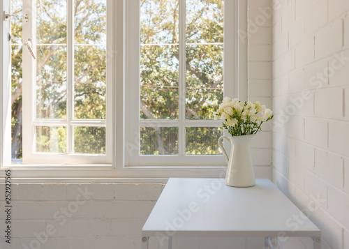 Fototapeta Naklejka Na Ścianę i Meble -  Closeup of white freesias in jug on table against window and painted brick wall (selective focus)