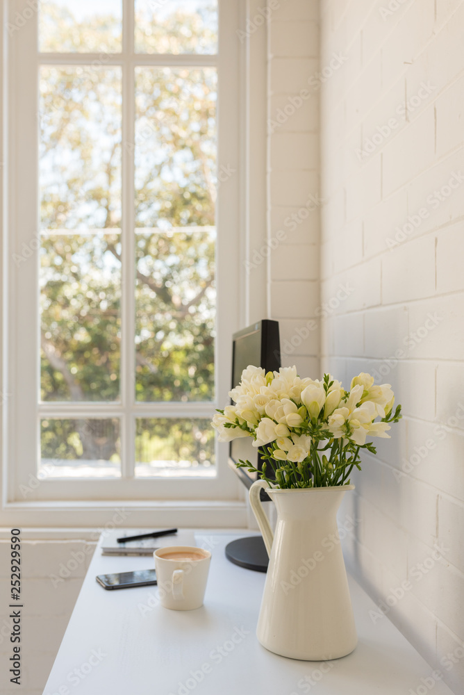 Vertical view of white freesias in jug on home office desk with computer and tea in background (selective focus) Vertical view of white freesias in jug on home office desk with computer and tea in background (selective focus)