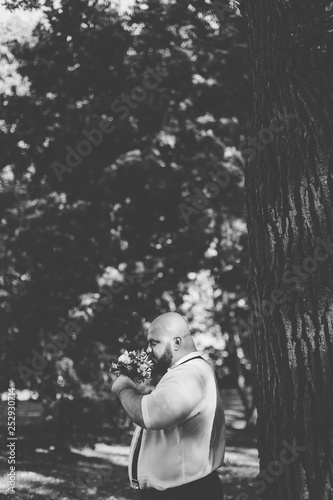 Stylish bearded fat groom with wedding bouquet in the park.