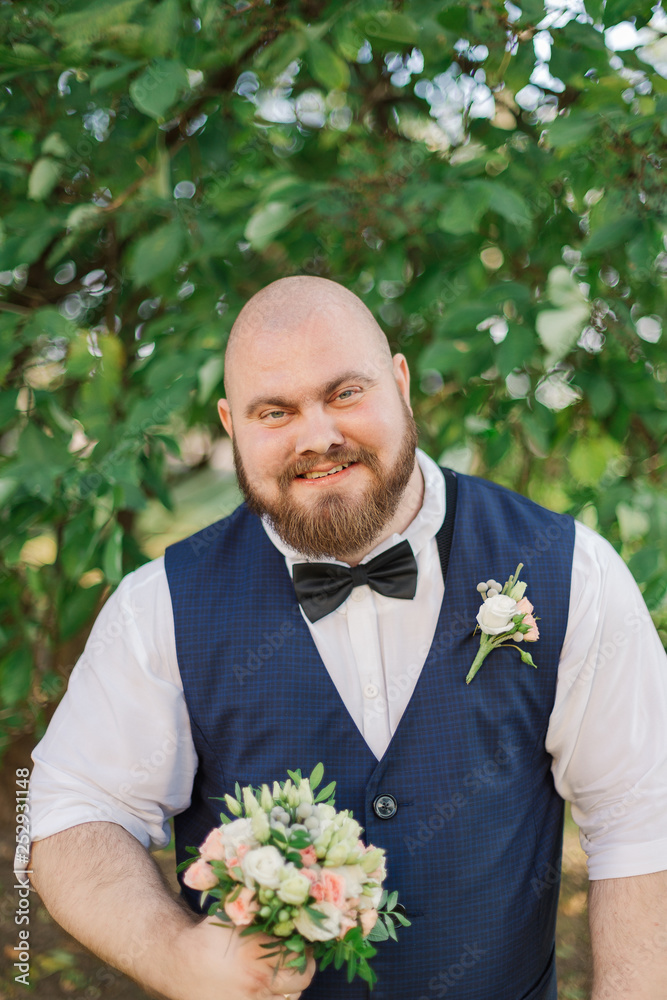 Stylish bearded fat groom with wedding bouquet in the park.