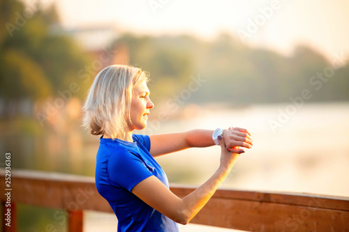 Photo from back sporty woman looks at clock on hand, standing on wooden bridge in park on summer. © nuclear_lily