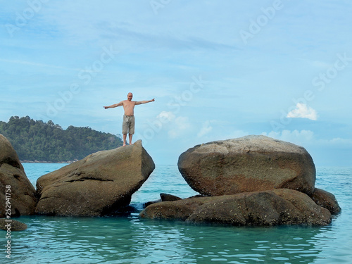 A man, alone, with a naked torso, in shorts, stands on a high stone in the sea with his arms spread to the sides