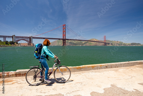 Young woman is sitting on a bicycle looking at the Golden Gate bridge from a pier in San Francisco, United States of America