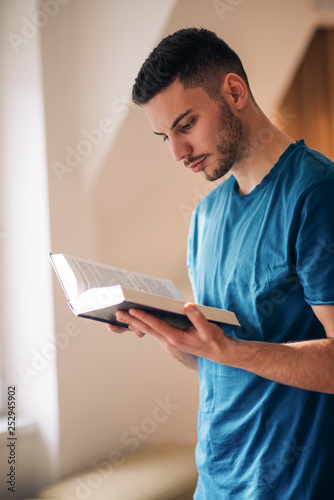 Student reading a book while standing and walking in the room next to the windows.