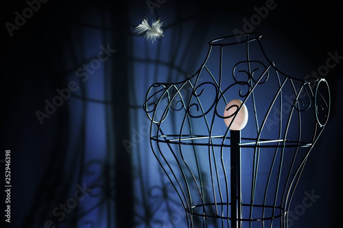 egg in a metal tailor doll like in a bird cage and a flying feather, surreal art concept for longing and loneliness, dark blue background with copy space