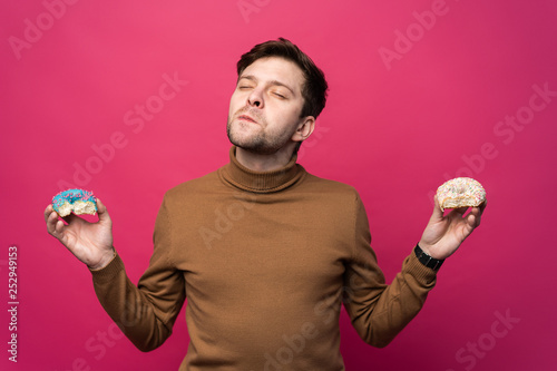 Displeased man holds two tasty ring donuts, feels unhappy as cant eat sweets, isolated over pink background. Confused stressed male.