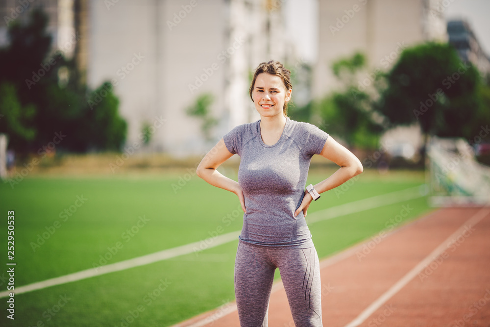 Portrait of a beautiful young caucasian woman with long hair in the tail and big breasts posing in gray sportswear standing training on a running stadium, a red rubber track in summer on a sunny day