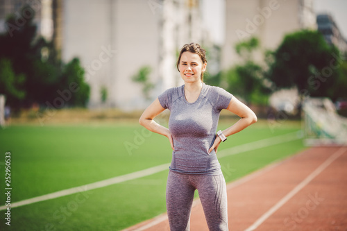 Portrait of a beautiful young caucasian woman with long hair in the tail  and big breasts posing in gray sportswear standing training on a running  stadium, a red rubber track in summer on a sunny day Stock Photo