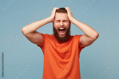 Man holding his head with his hands shouts loudly, facial expression of anger, isolated over blue background.Guy with closed eyes wide opened mouth screams from pain hopelessness despair