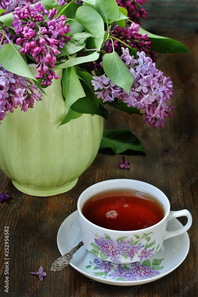 Tea and a bouquet of lilac. Tea in a beautiful cup and a bouquet of lilac in a vase. A spring tea-party. Soft focus Tea and a bouquet of lilac. Tea in a beautiful cup and a bouquet of lilac in a vase. A spring tea-party. Soft focus